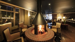A cozy indoor seating area with a central fire feature, comfortable chairs, and a view through large windows.
