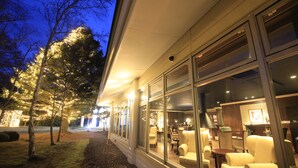 A building with large windows, a seating area with chairs and tables, and trees with lights.