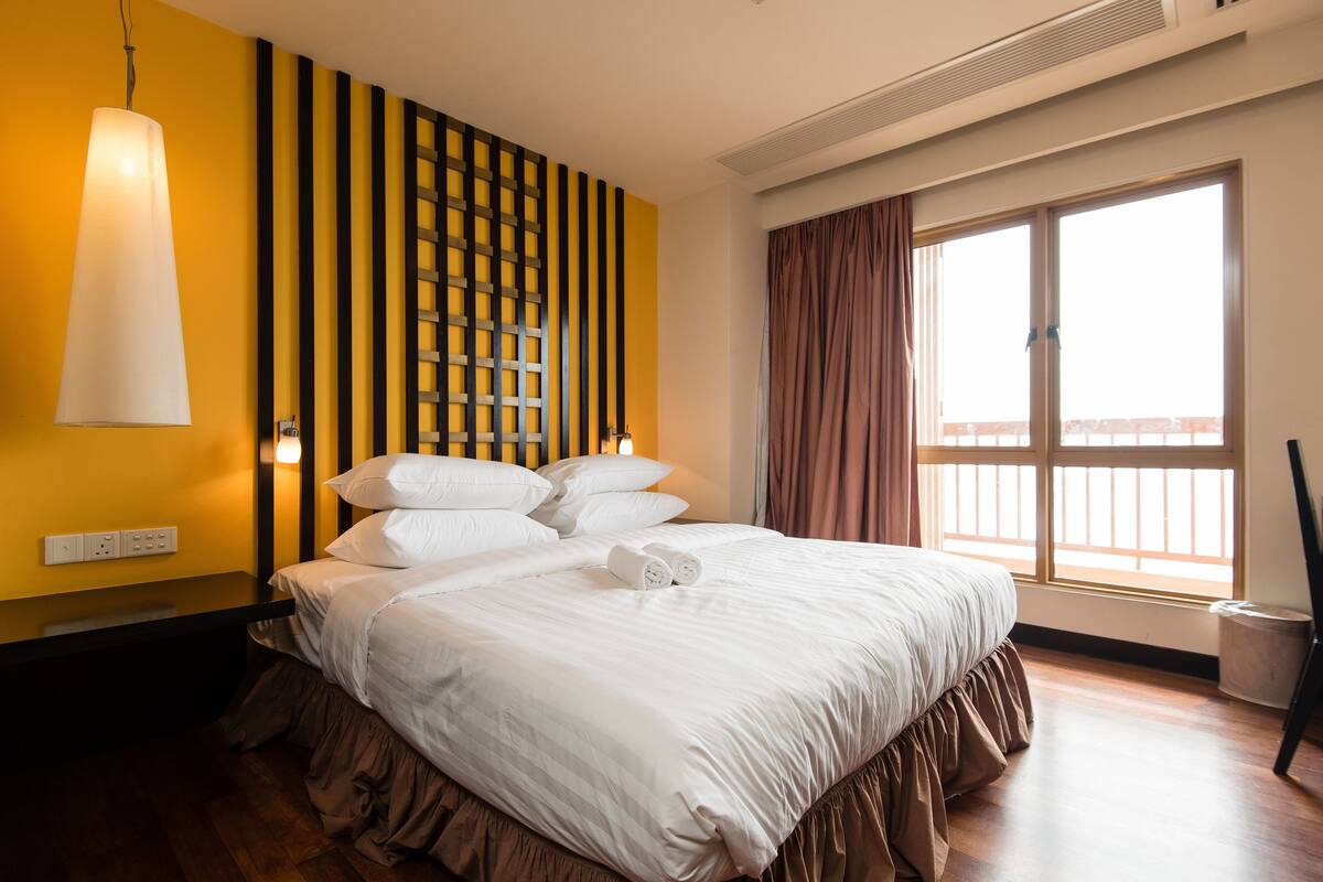 A hotel room with a large bed, a wooden floor, a window with curtains, and a wall with vertical stripes.