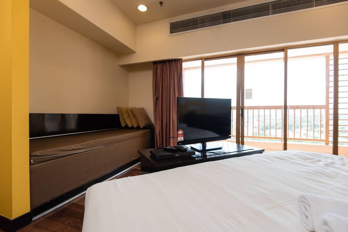 A hotel room with a bed, a television on a stand, a sofa, and a large window with a view.