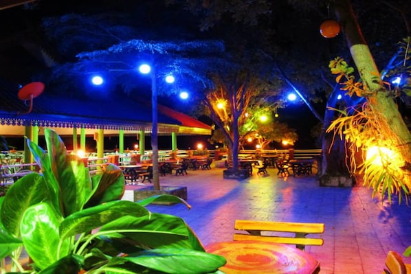 An outdoor dining area with tables and chairs, illuminated by overhead lights.