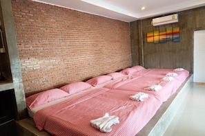 A long, narrow hotel room with a brick wall, a large bed with pink bedding, and a colorful artwork on the wall.