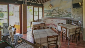 A rustic dining area with wooden tables and chairs, a brick wall with a mural, and a tiled floor. - Villa Maria (Vicopisano)