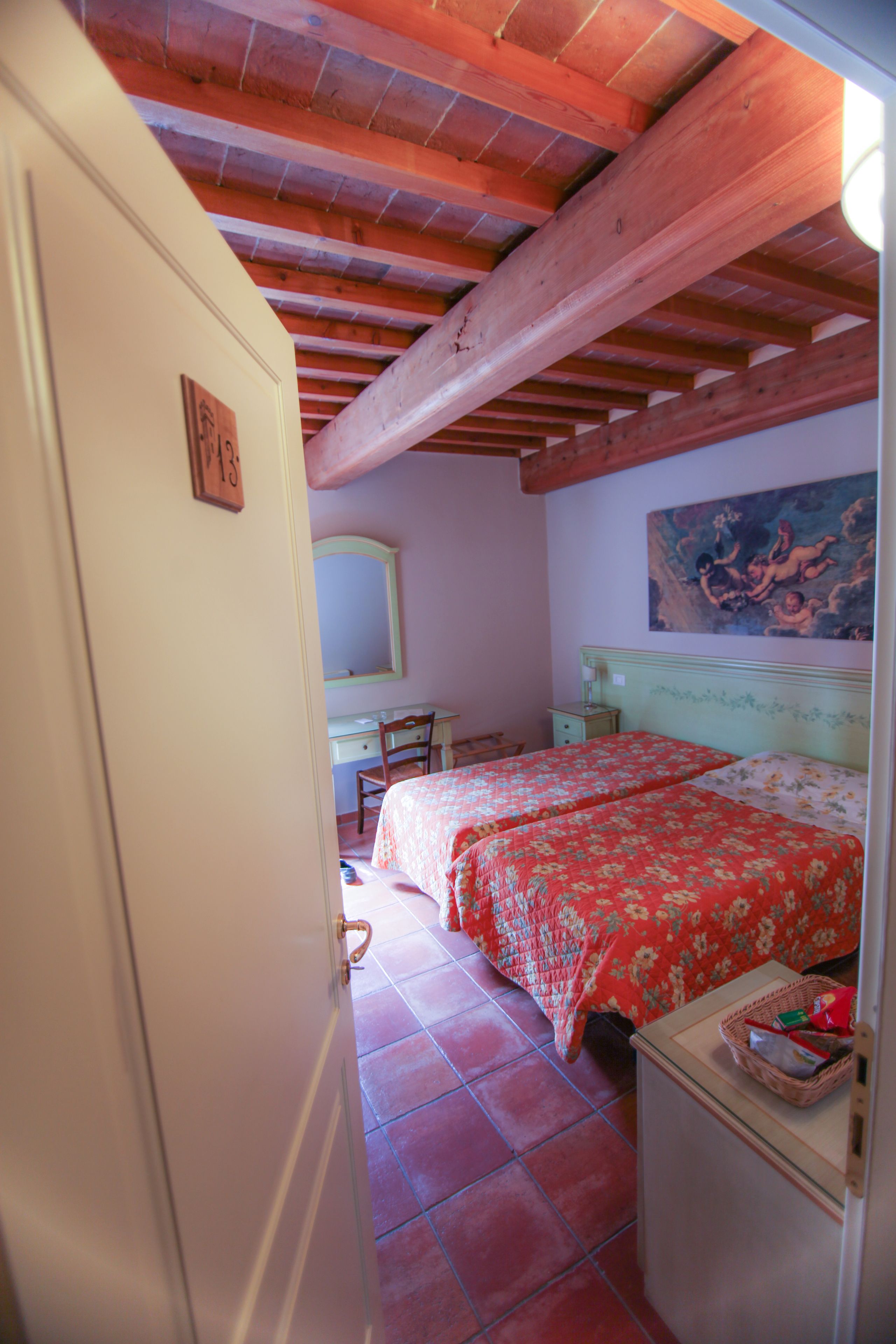 Un dormitorio con techo de madera, cama con ropa de cama floral, escritorio con silla, un espejo y un cuadro en la pared.