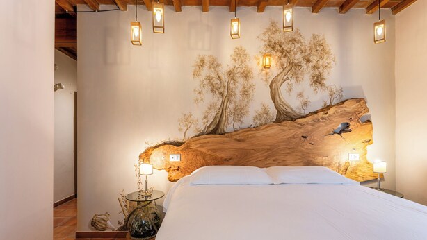 A bedroom with a large bed, a wooden headboard, and a wall mural of trees. There are bedside lamps and a small table with a lamp. - Villa Maria (Vicopisano)