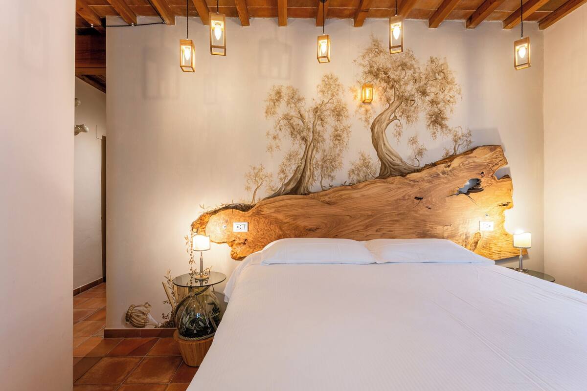 A bedroom with a large bed, a wooden headboard, and a wall mural of trees. There are bedside lamps and a small table with a lamp.