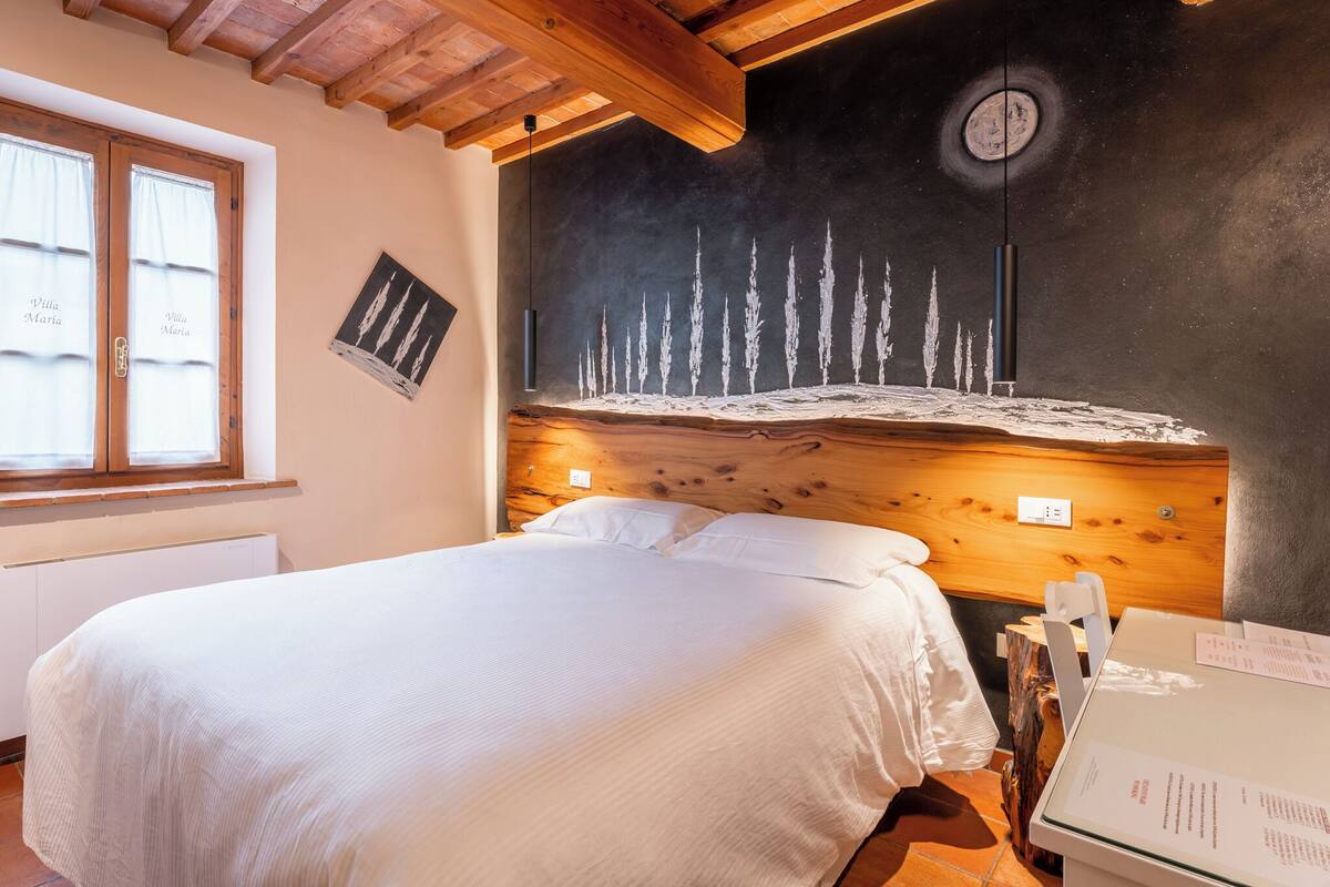 A bedroom with a bed, a window with curtains, a wall painting, and a wooden ceiling.