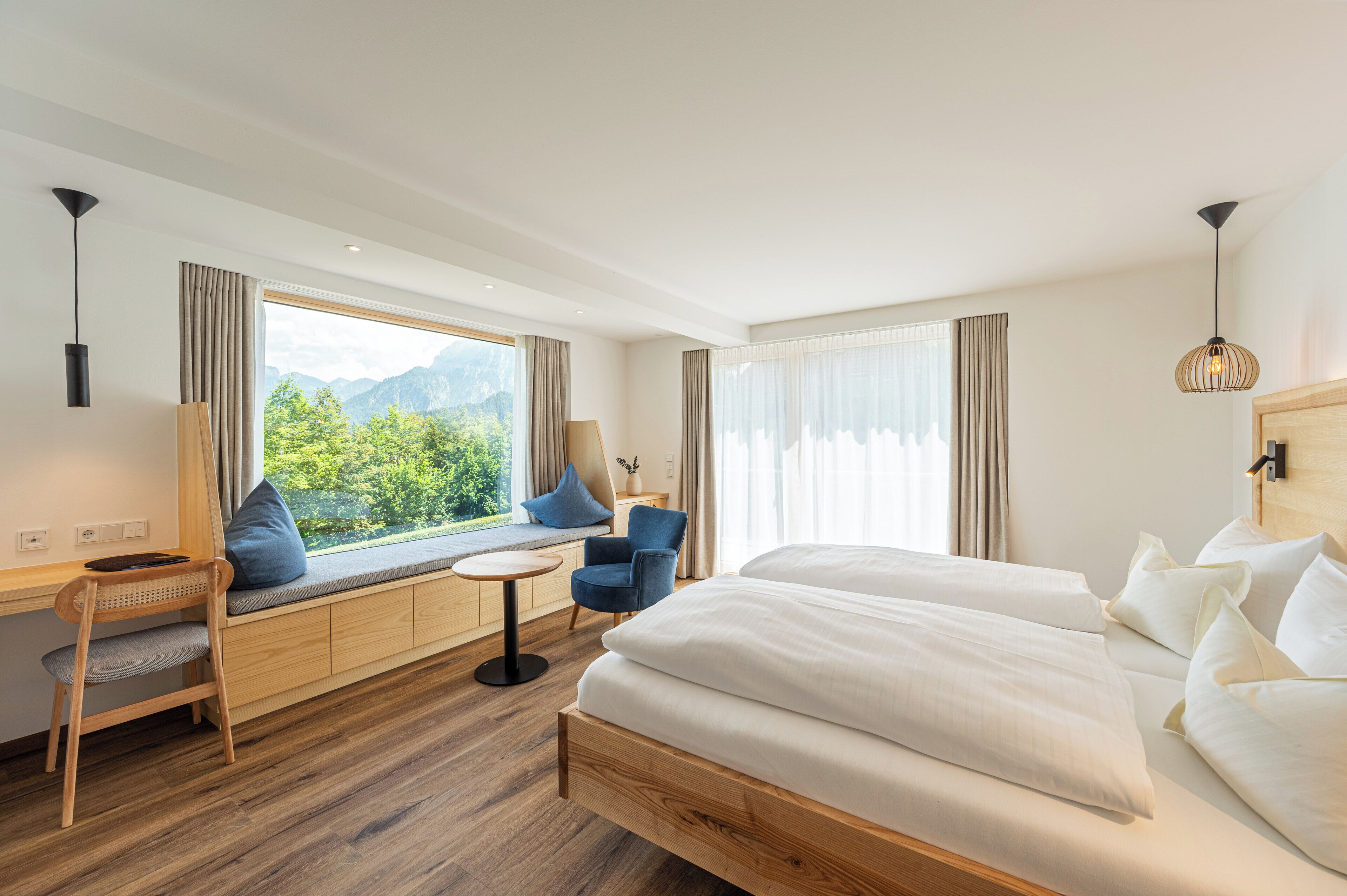 Habitación de hotel con una cama grande, un escritorio con silla, una mesita y vistas a montañas a través de la ventana.