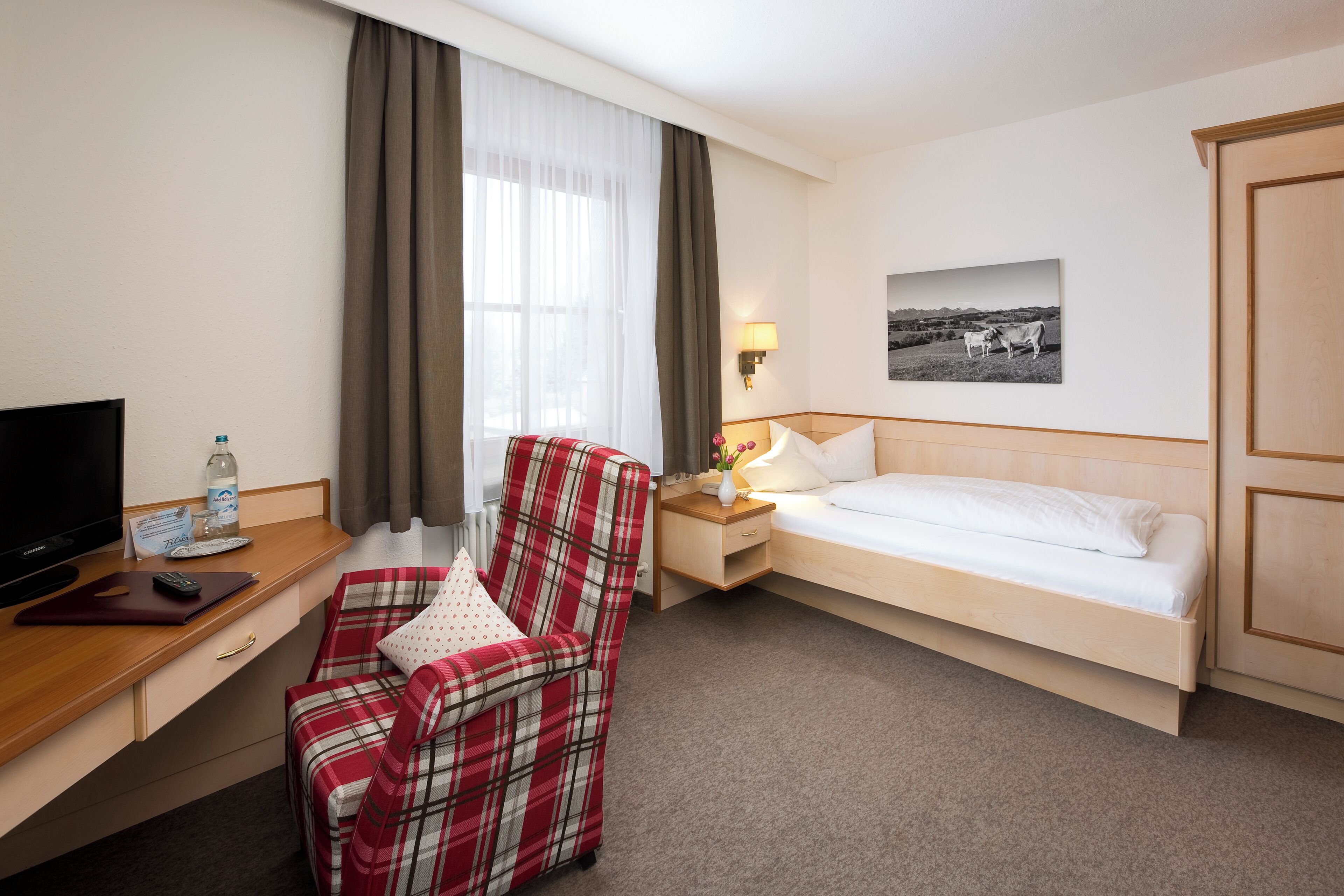 A hotel room with a bed, a desk with a TV, a chair, a nightstand with a lamp, and a picture on the wall.