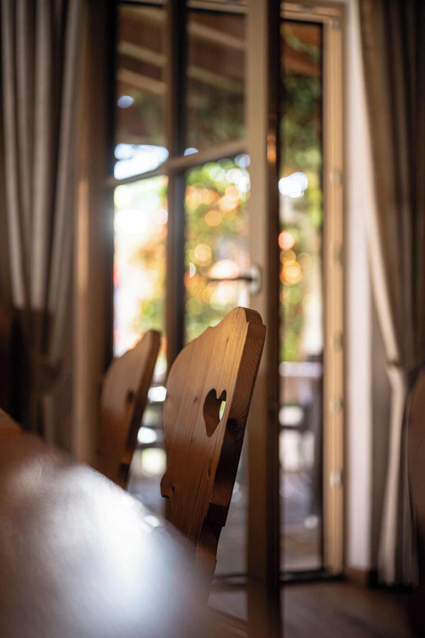 A wooden chair with a heart-shaped cutout, a wooden table, and a view of the outdoors through a glass door.
