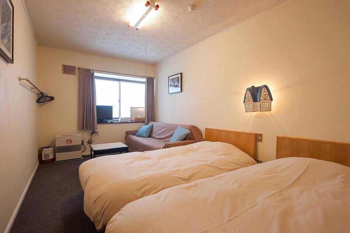 A hotel room with two beds, a desk, a chair, a window with curtains, a wall-mounted heater, and a wall-mounted lamp.