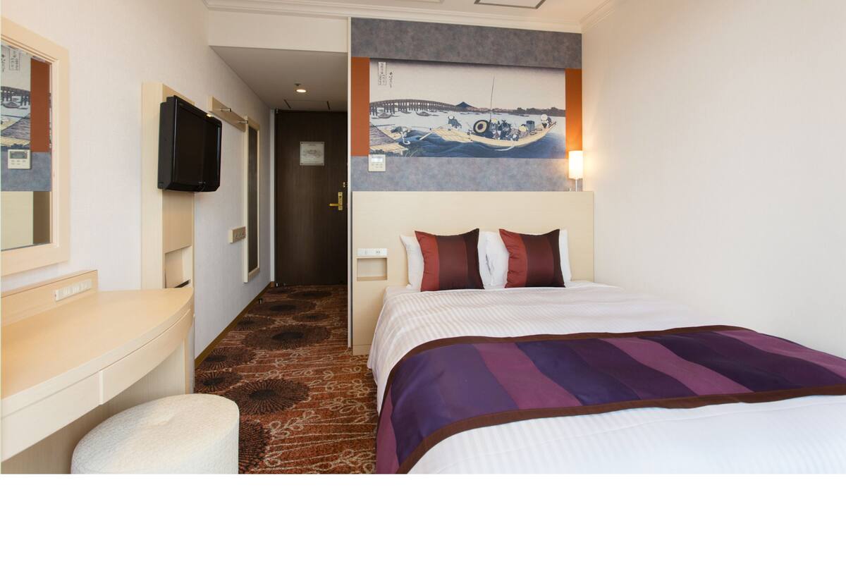 A hotel room with a bed, a desk, a television, and a painting on the wall.