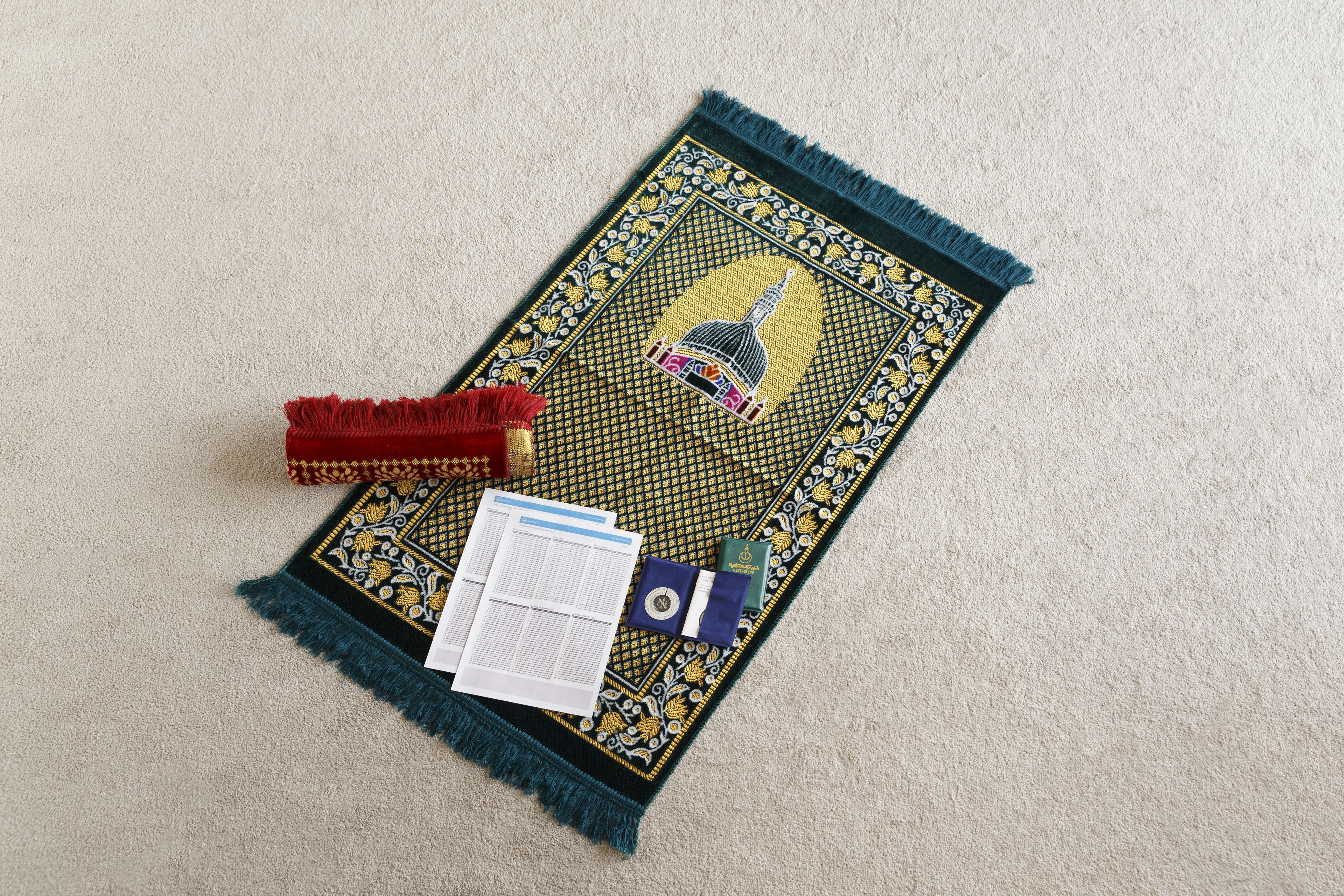 A prayer mat with a central emblem, a red tassel, and a booklet on a carpeted floor.
