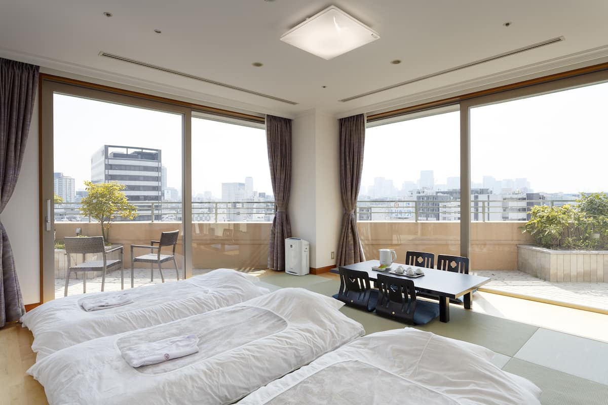 A hotel room with two beds, a dining table, chairs, and large windows offering a city view.