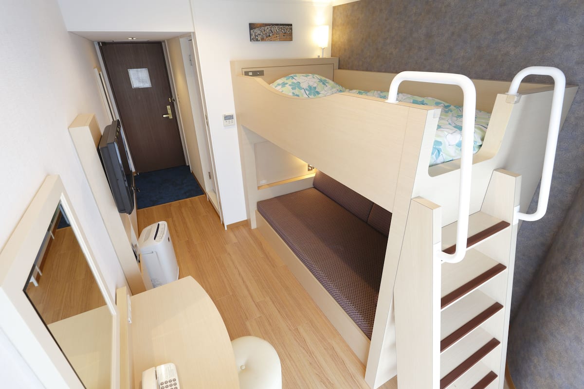 A bunk bed room with a desk and chair, a door, and a small trash bin.