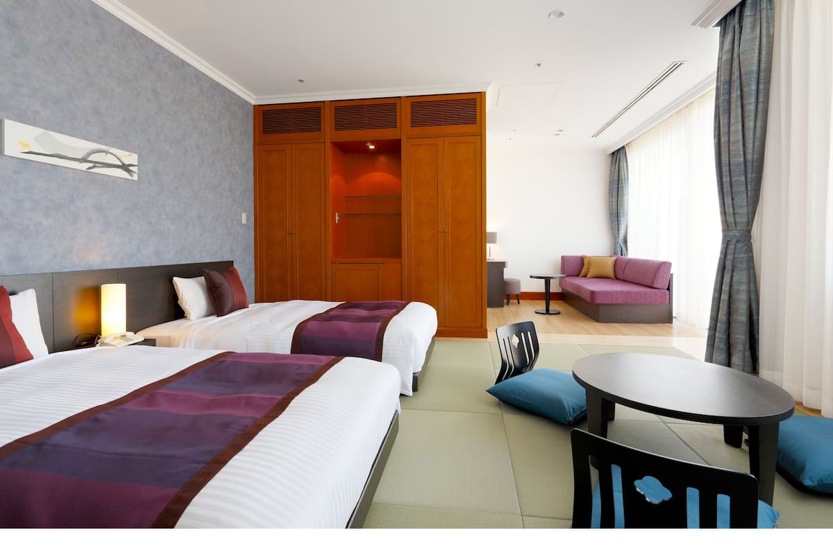 A hotel room with two beds, a sofa, a small table, and a view of the outdoors.