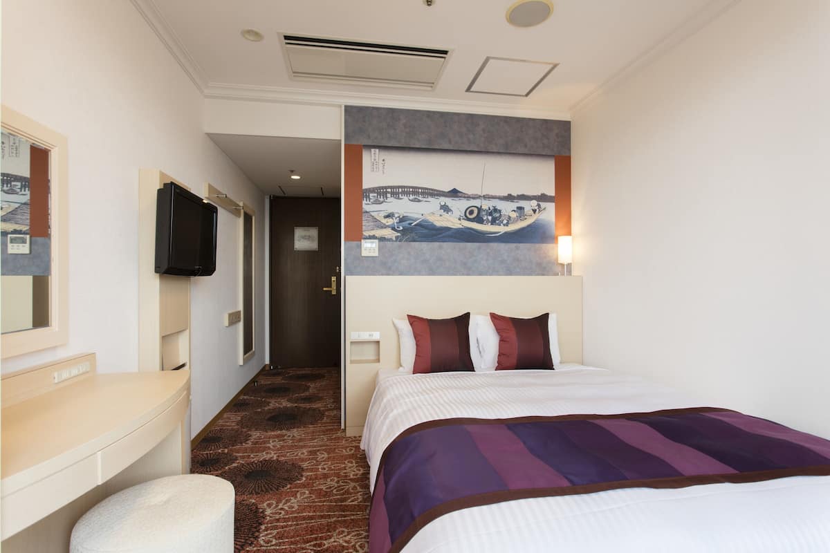 A hotel room with a bed, a TV, a painting on the wall, and a carpeted floor.