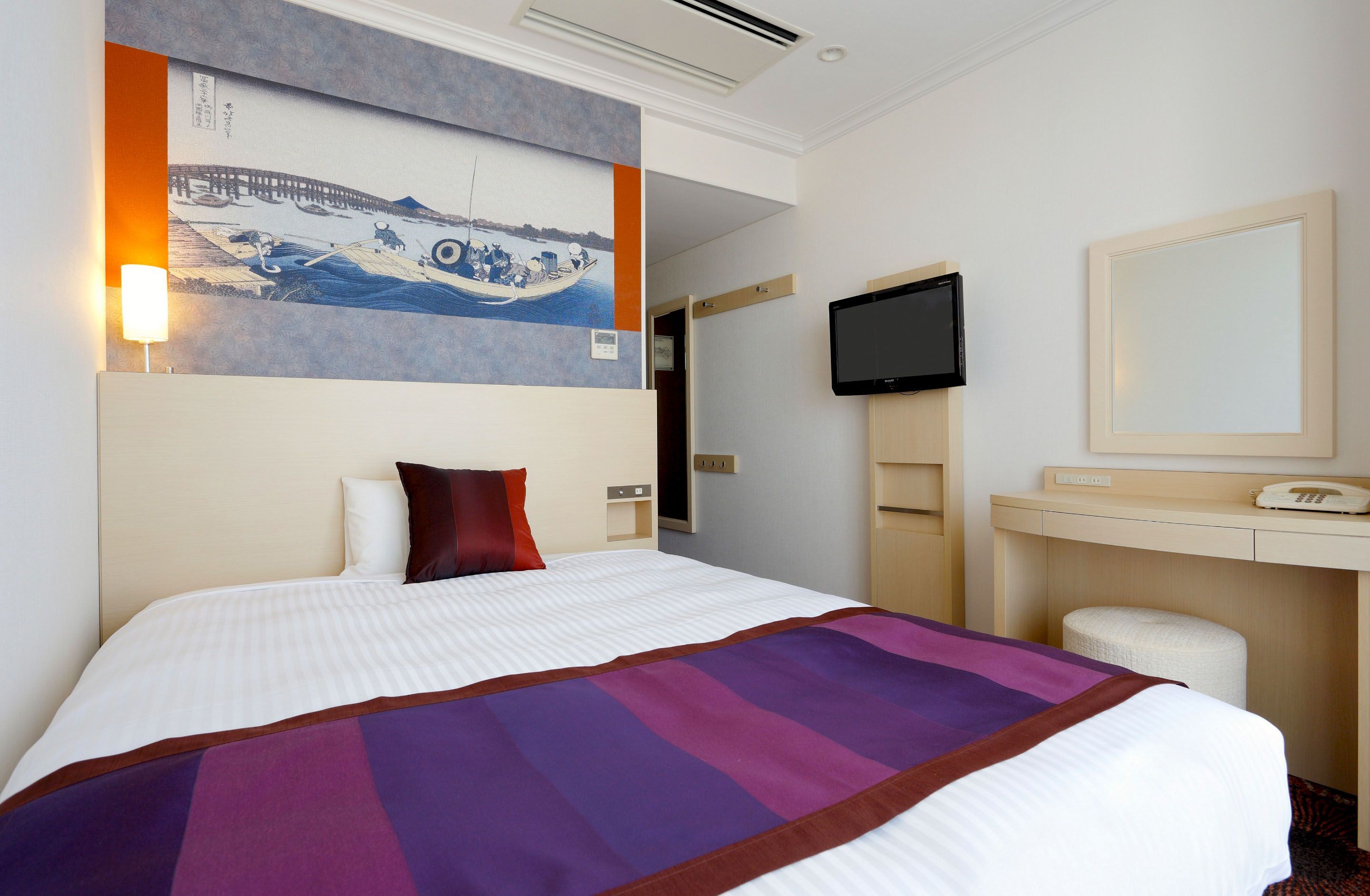 A hotel room with a bed, a desk, a television, and a painting on the wall.