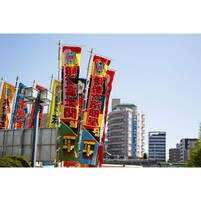 Egy sor színes, japán feliratokkal díszített zászló, a háttérben egy modern épület, és tiszta, kék égbolt.