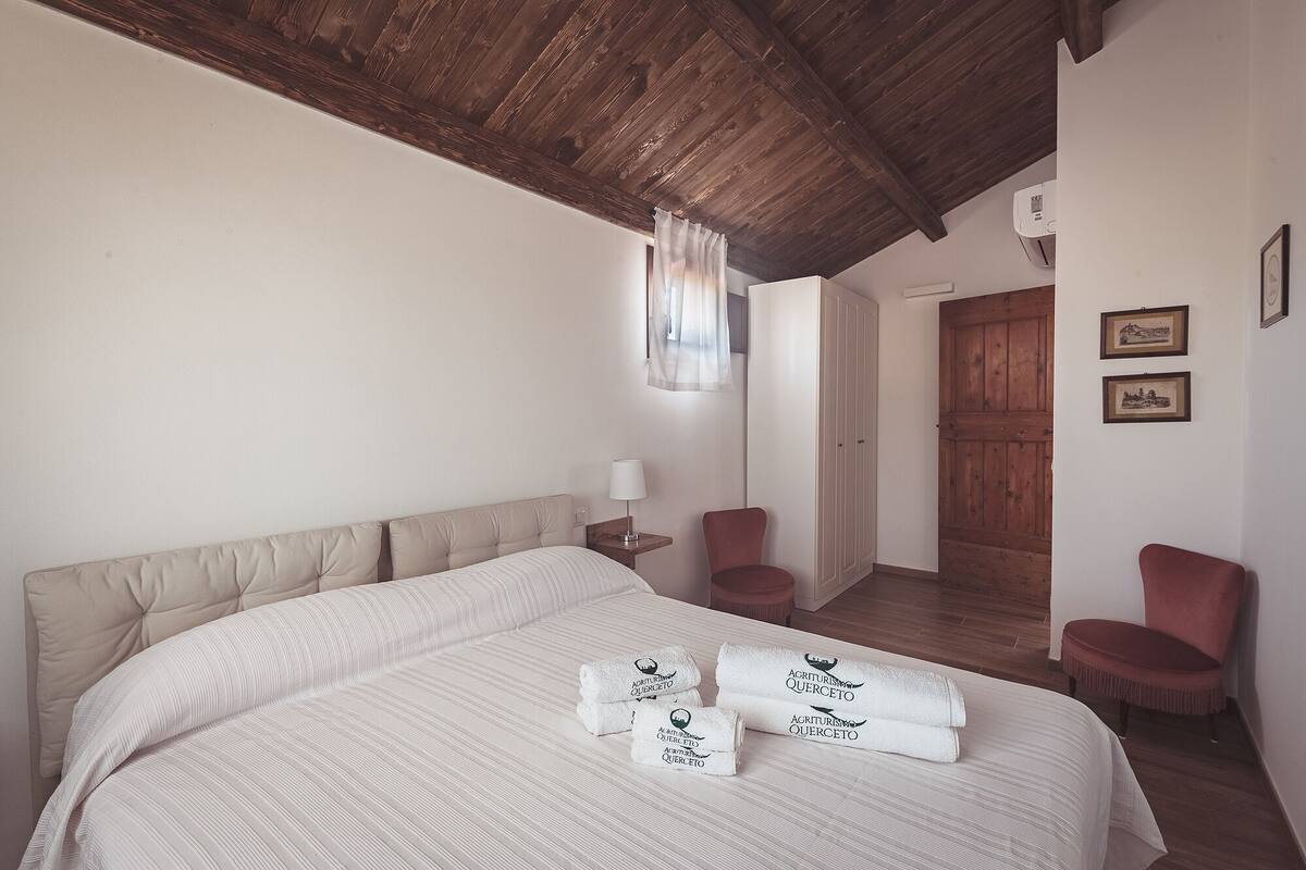 A bedroom with a large bed, two chairs, a wooden wardrobe, and a door leading to another room.