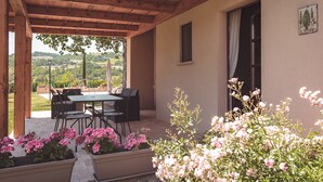 Een terras buiten met een houten pergola, een tafel met stoelen en potten met roze bloemen.