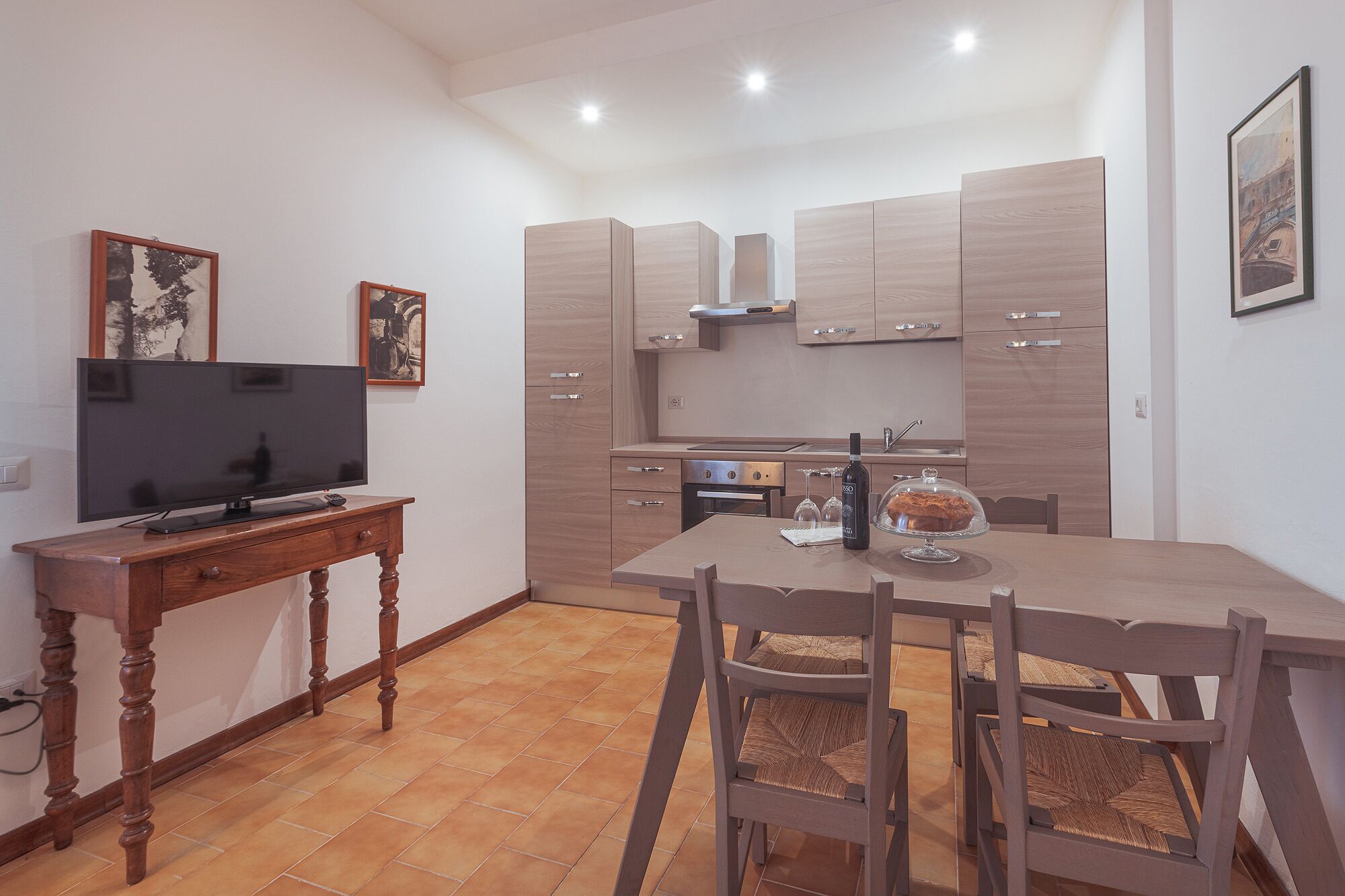 A modern kitchen with wooden cabinets, a dining table with chairs, a television, and artwork on the walls.