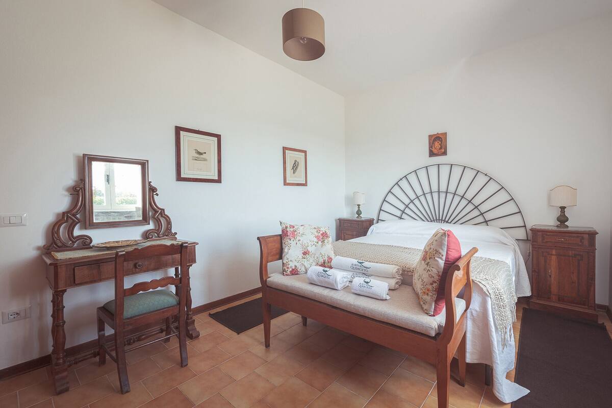 A bedroom with a bed, a wooden desk, a chair, a mirror, and framed pictures on the wall.