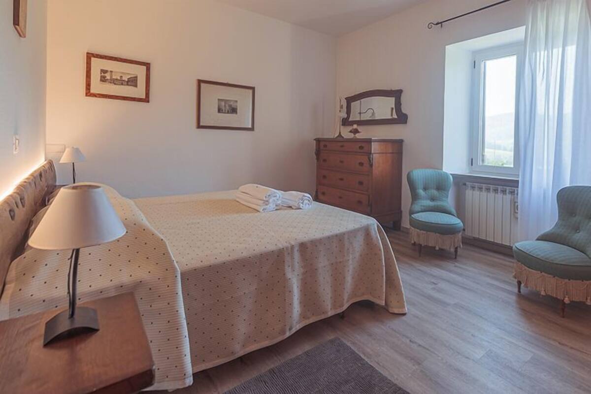 A bedroom with a bed, a dresser, two chairs, a lamp, and a window with curtains.