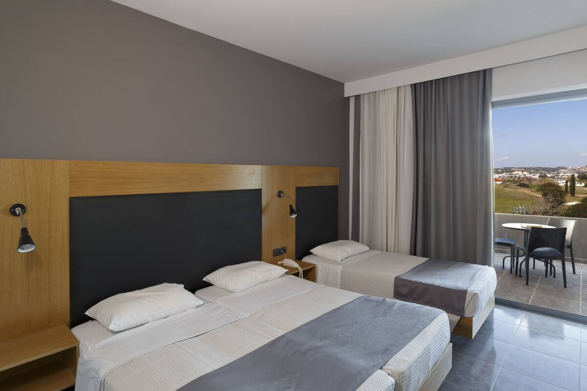 Quarto de hotel com duas camas, uma varanda com mesa e cadeiras, e vista para a cidade.