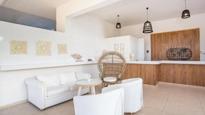 A modern, well-lit living area with a white sofa, two white armchairs, a wicker chair, and a small round table. There are three pendant lights hanging from the ceiling, and a wooden accent wall with a world map design. - Evita Resort (Rhodes)
