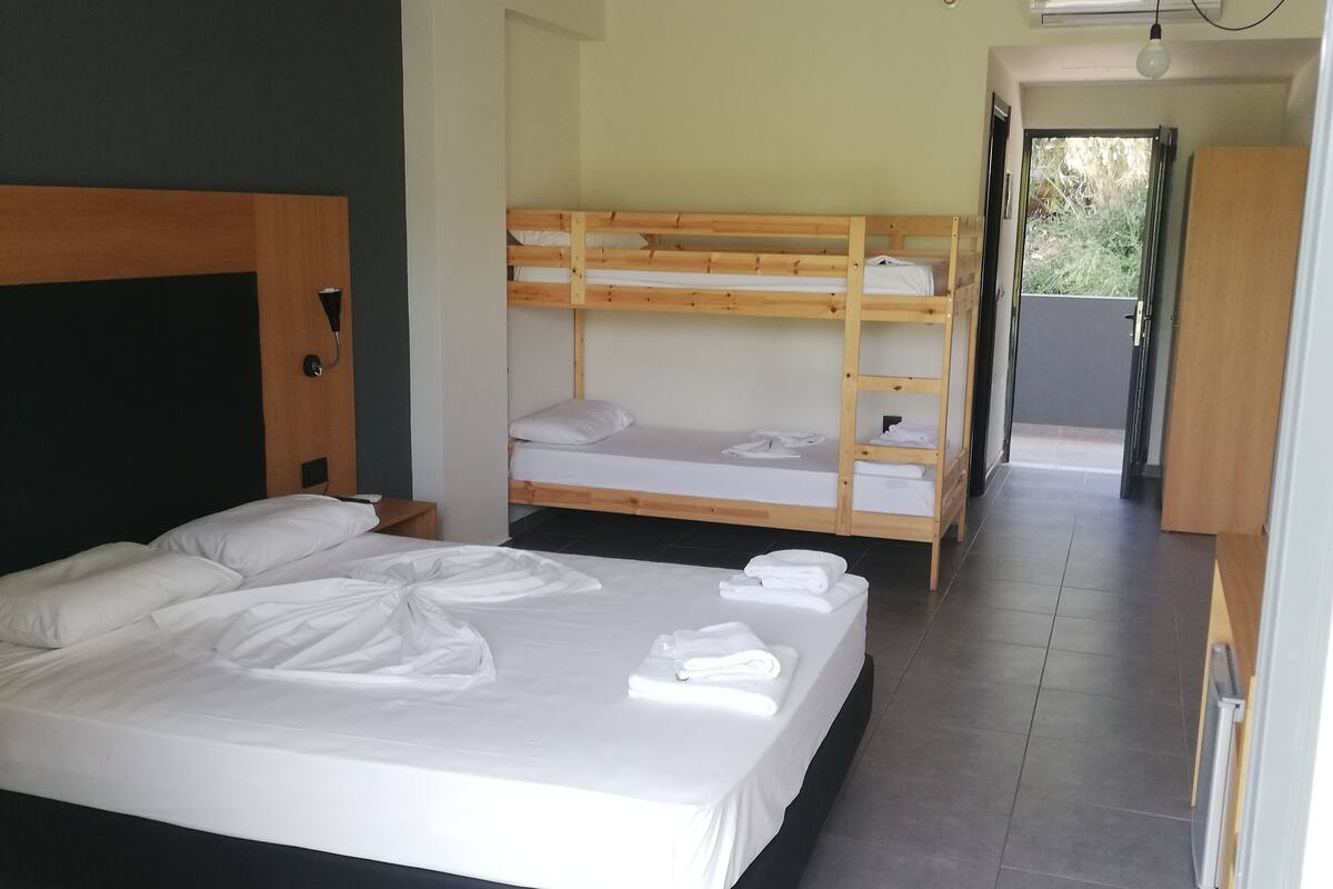 Quarto de hotel moderno com uma cama grande, uma beliche e uma kitchenette pequena.