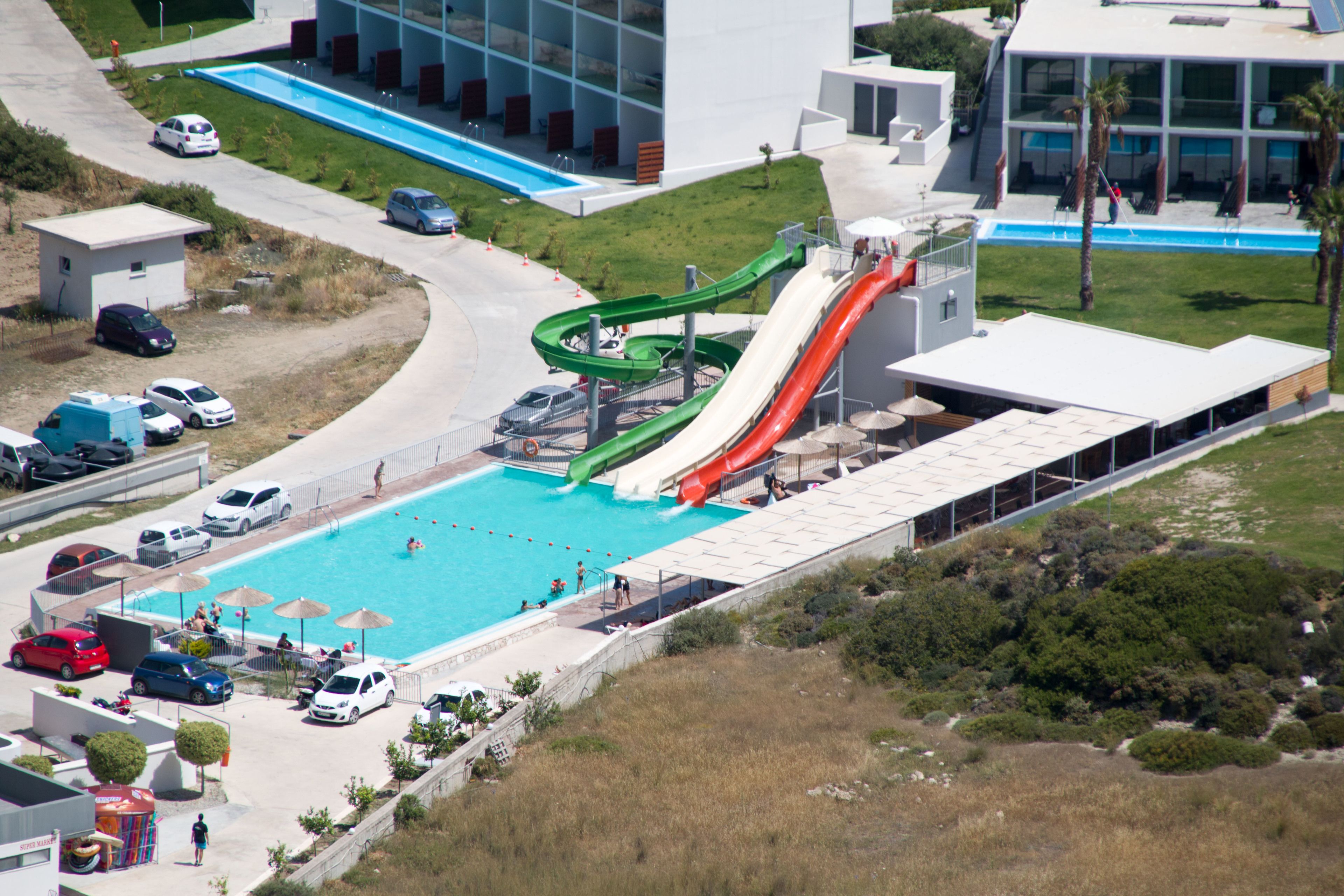 Vista aerea di una piscina con due scivoli, circondata da auto parcheggiate e moderni edifici.
