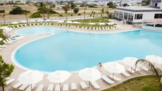 A large swimming pool with sun loungers and umbrellas, surrounded by a landscaped area with a view of distant buildings. - Evita Resort (Rhodes)