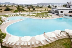 A large swimming pool with sun loungers and umbrellas, surrounded by a landscaped area with a view of distant buildings. - Evita Resort (Rhodes)