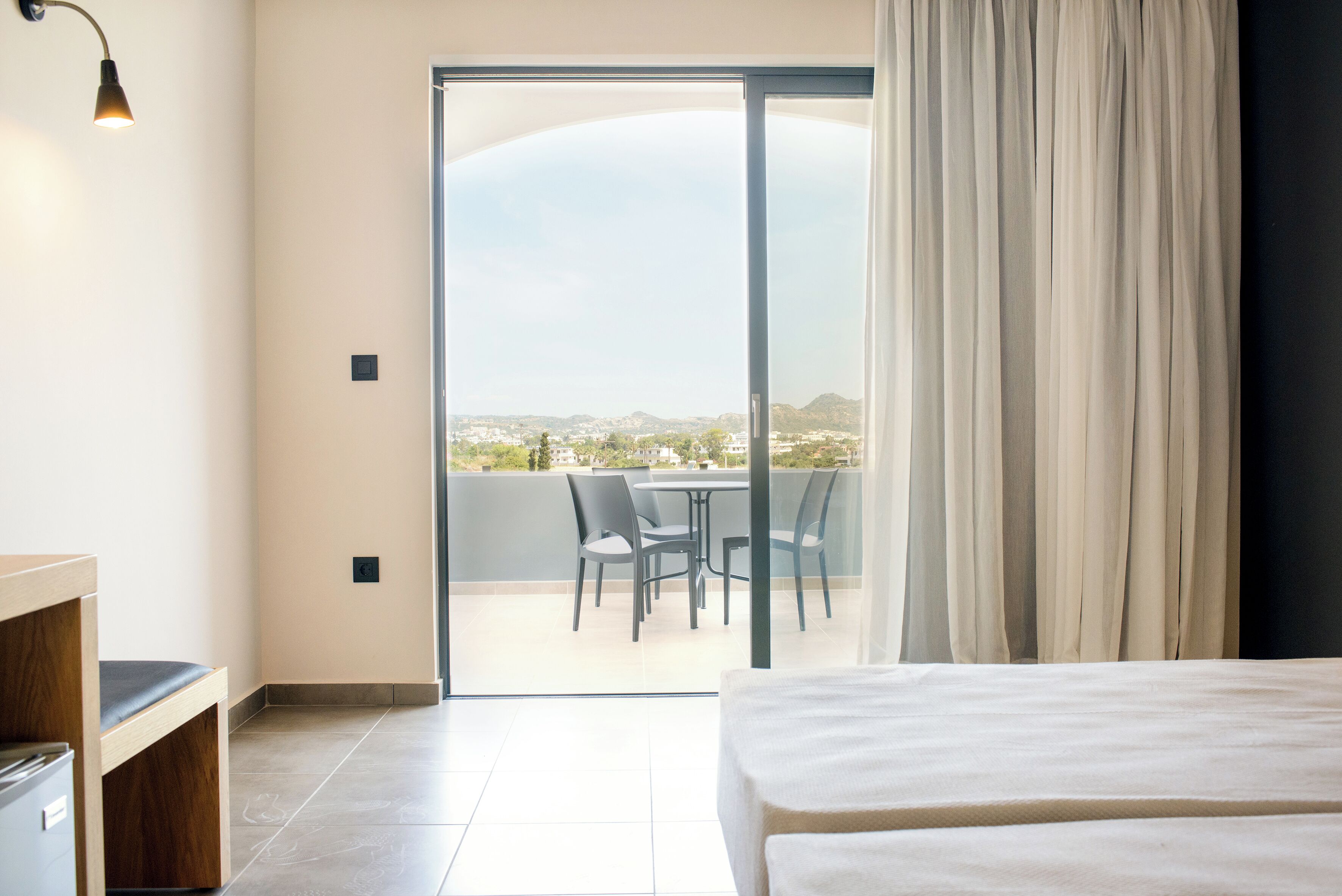 Habitación de hotel con cama, escritorio, silla, mesa y vista a un paisaje urbano a través de una puerta corredera de vidrio.