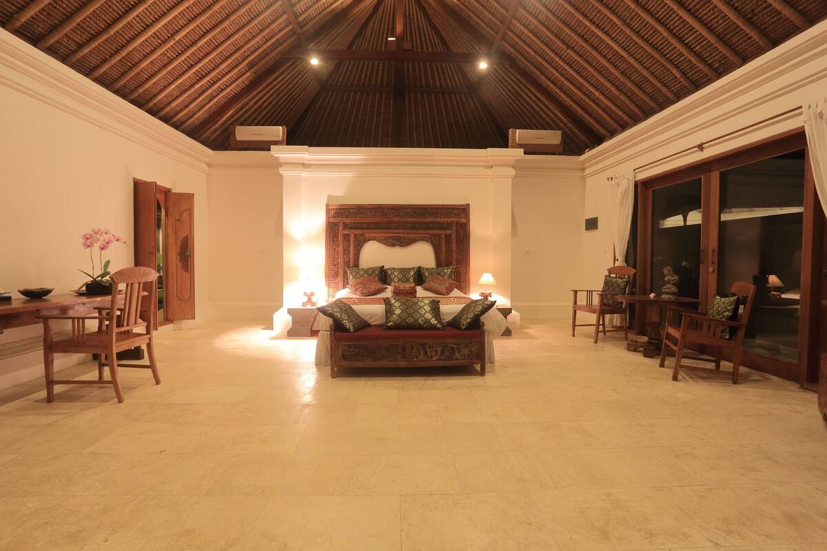 A spacious room with a high ceiling, a bed with ornate headboard, and a seating area with chairs and a sofa.