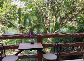 Un área de estar al aire libre con una mesa de madera y dos sillas, rodeada de abundante vegetación y vistas a un cuerpo de agua.
