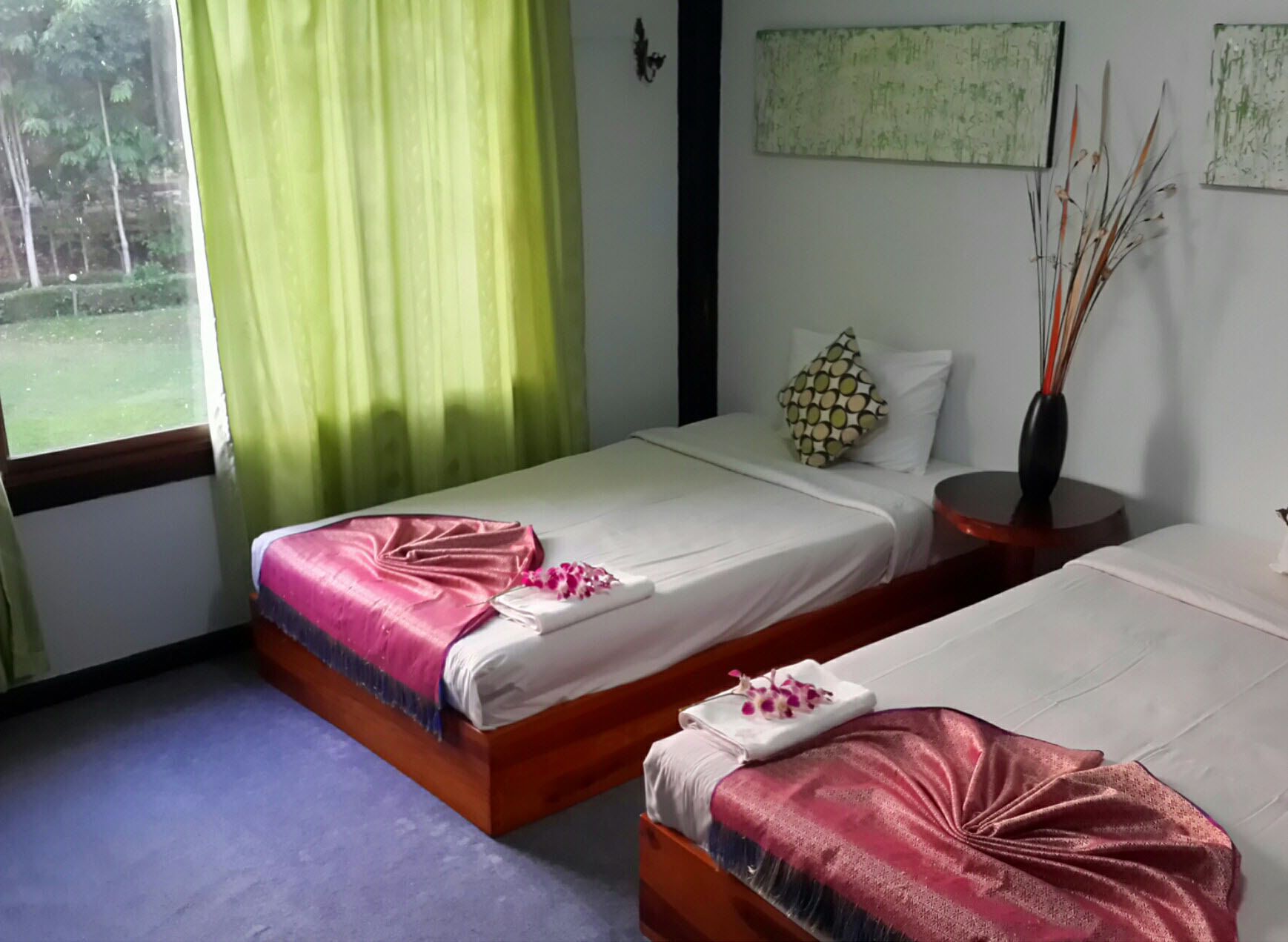 Habitación de hotel con dos camas, una ventana con cortinas verdes, un jarrón con plantas secas y un cuadro en la pared.