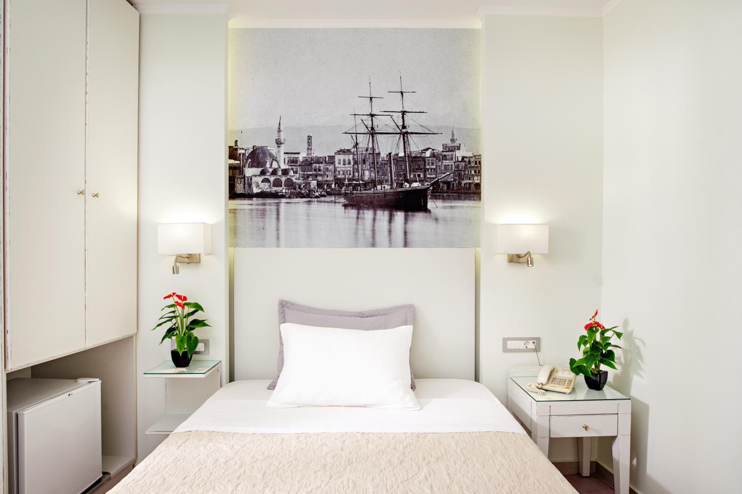 A hotel room with a bed, a nightstand with a lamp, a small table with a phone, and a wall-mounted picture of a harbor scene.