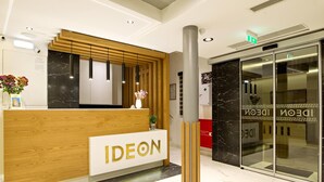 A reception desk with the word 'IDEON' prominently displayed, a modern interior design with wooden accents and marble flooring, and a glass elevator door with the same branding. - Ideon Hotel (Chania)