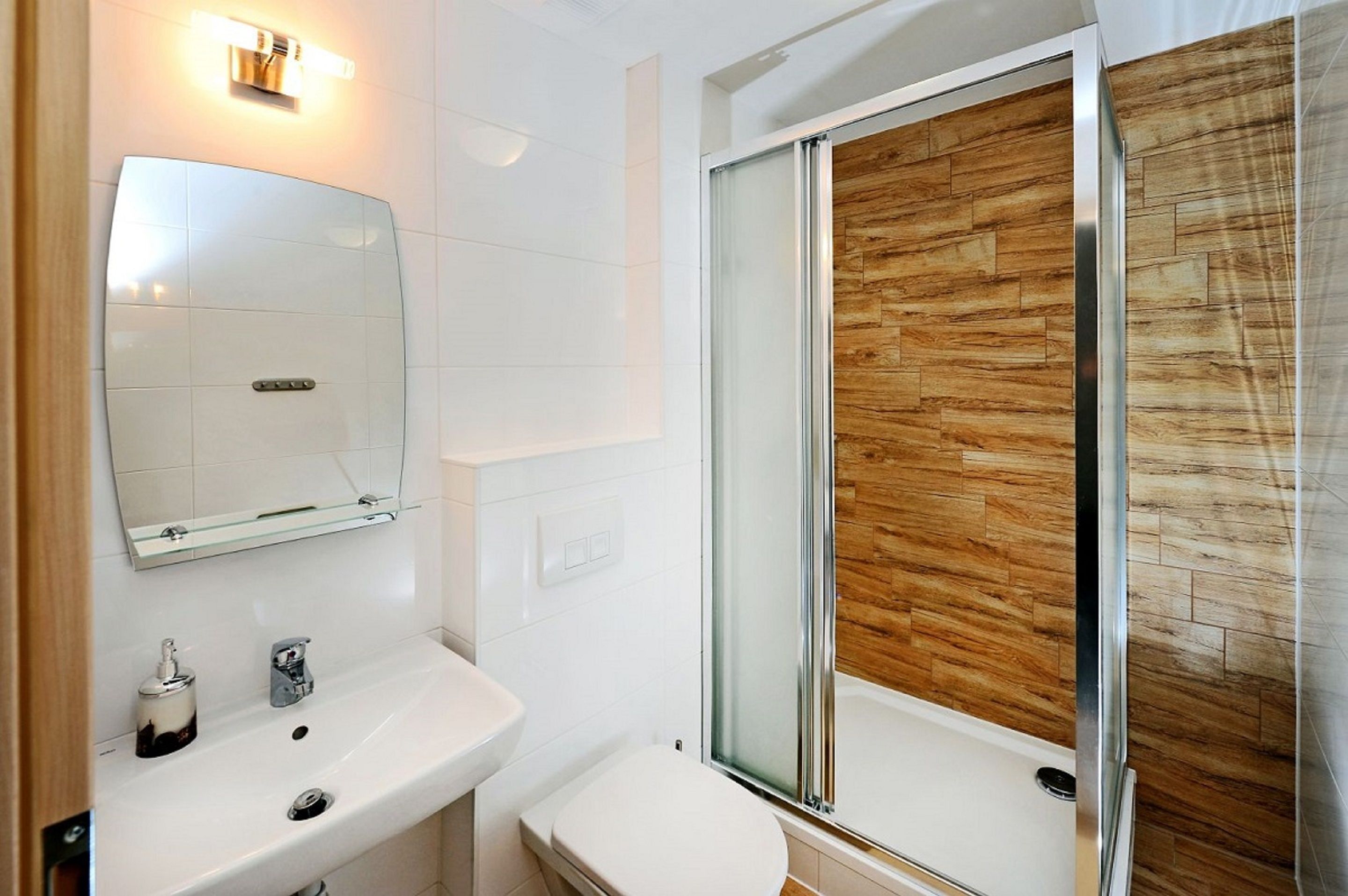 A modern bathroom with a white sink, mirror, and toilet. It features a glass shower enclosure with a wooden wall panel.