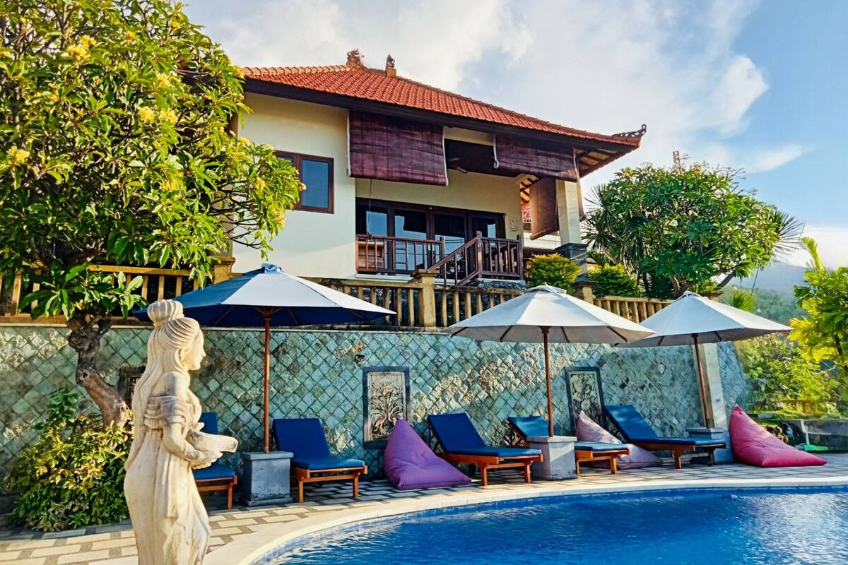 A poolside area with lounge chairs and umbrellas, a statue, and a building with a tiled facade and balcony.