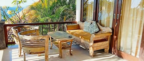A wooden outdoor seating area with a table and chairs, a sofa with cushions, and a view of lush greenery and mountains in the background.