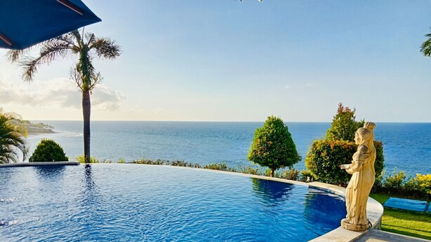 Piscina con vista al océano, rodeada de palmeras y una estatua.
