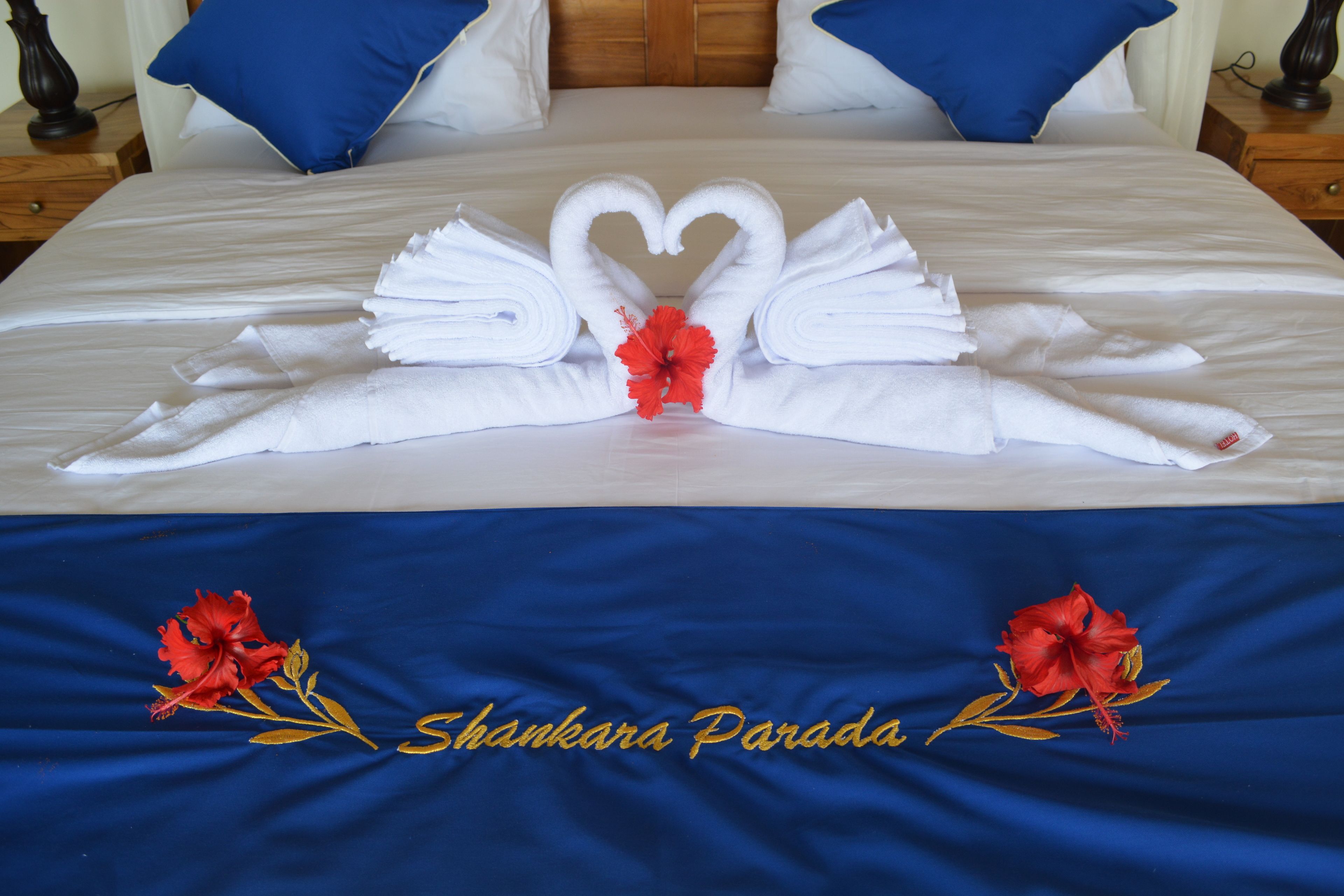 Un lit avec du linge de lit blanc, une couvre-lit bleue ornée d’une broderie représentant « Shankara Parada », et un arrangement floral central composé d’une fleur d’hibiscus rouge.
