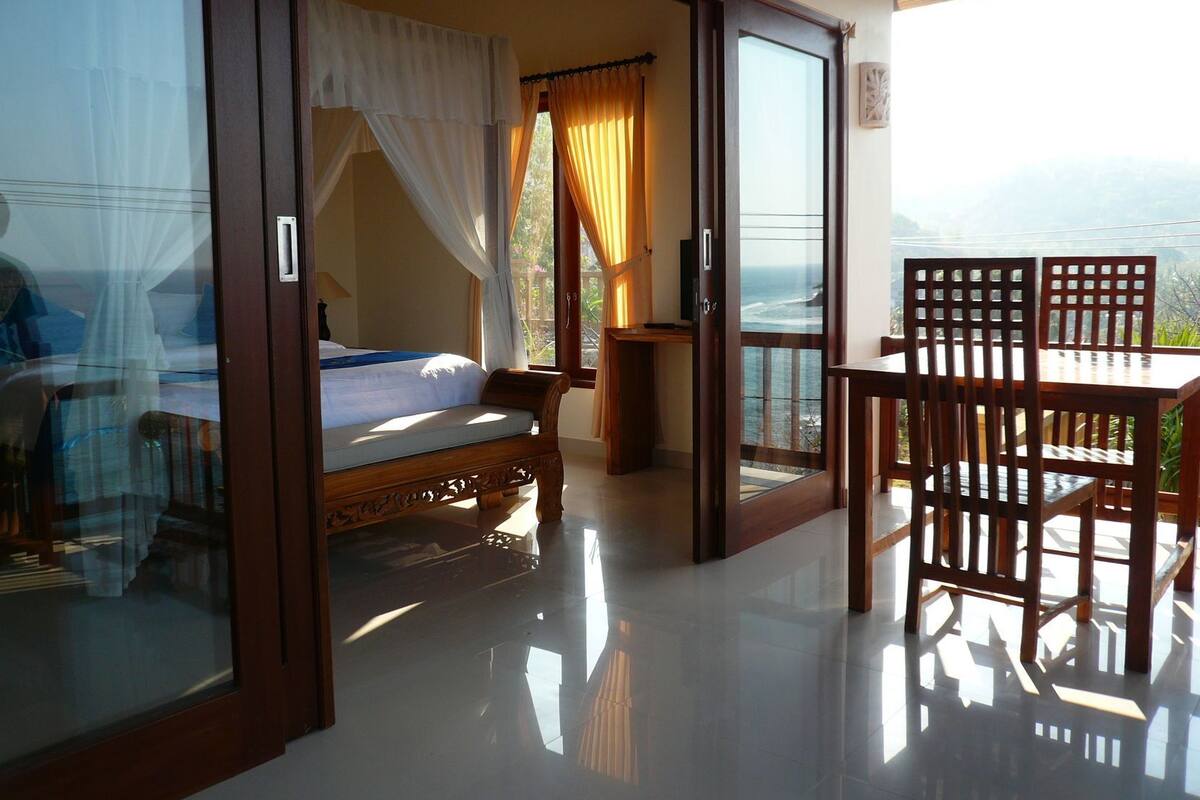 A room with a bed, a table with chairs, and a balcony with a view.