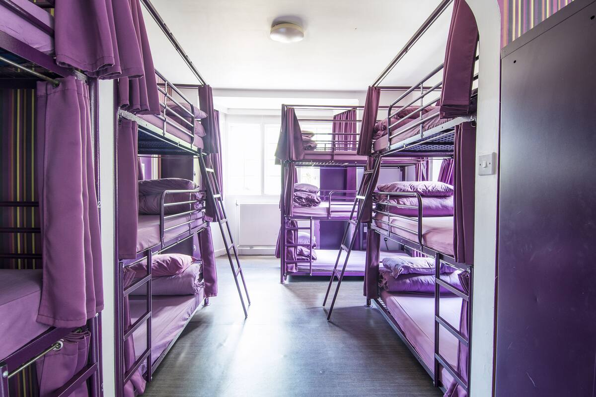 A dormitory with bunk beds arranged in a corridor, with purple curtains and a window at the end.