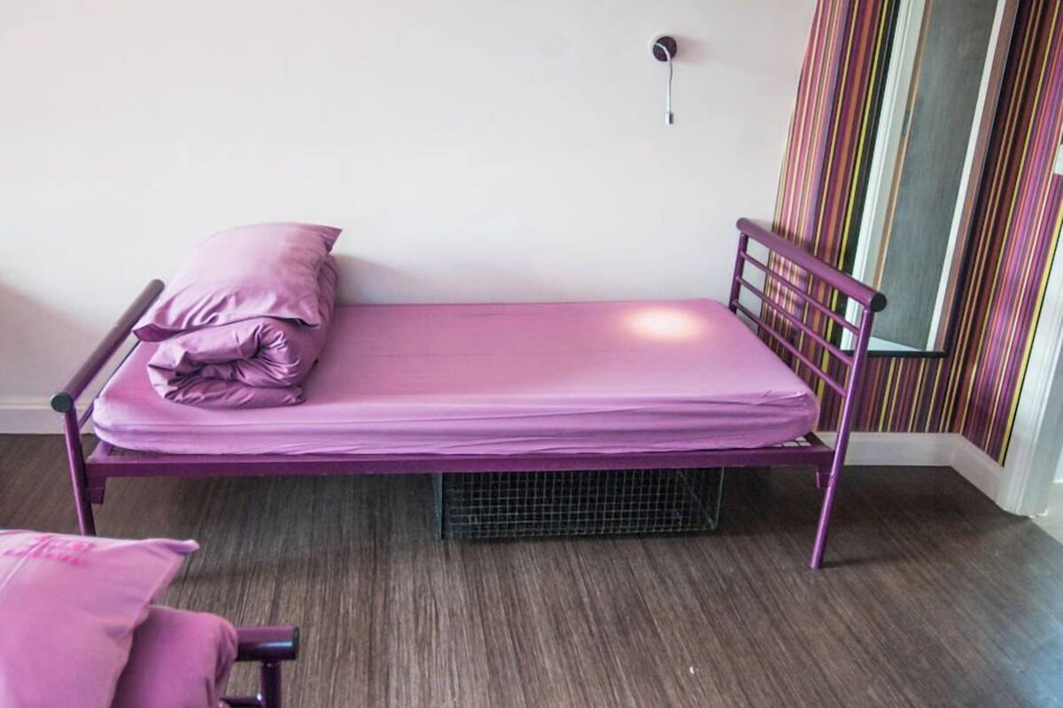 A single bed with a metal frame and a purple mattress in a room with a wooden floor and a striped curtain.