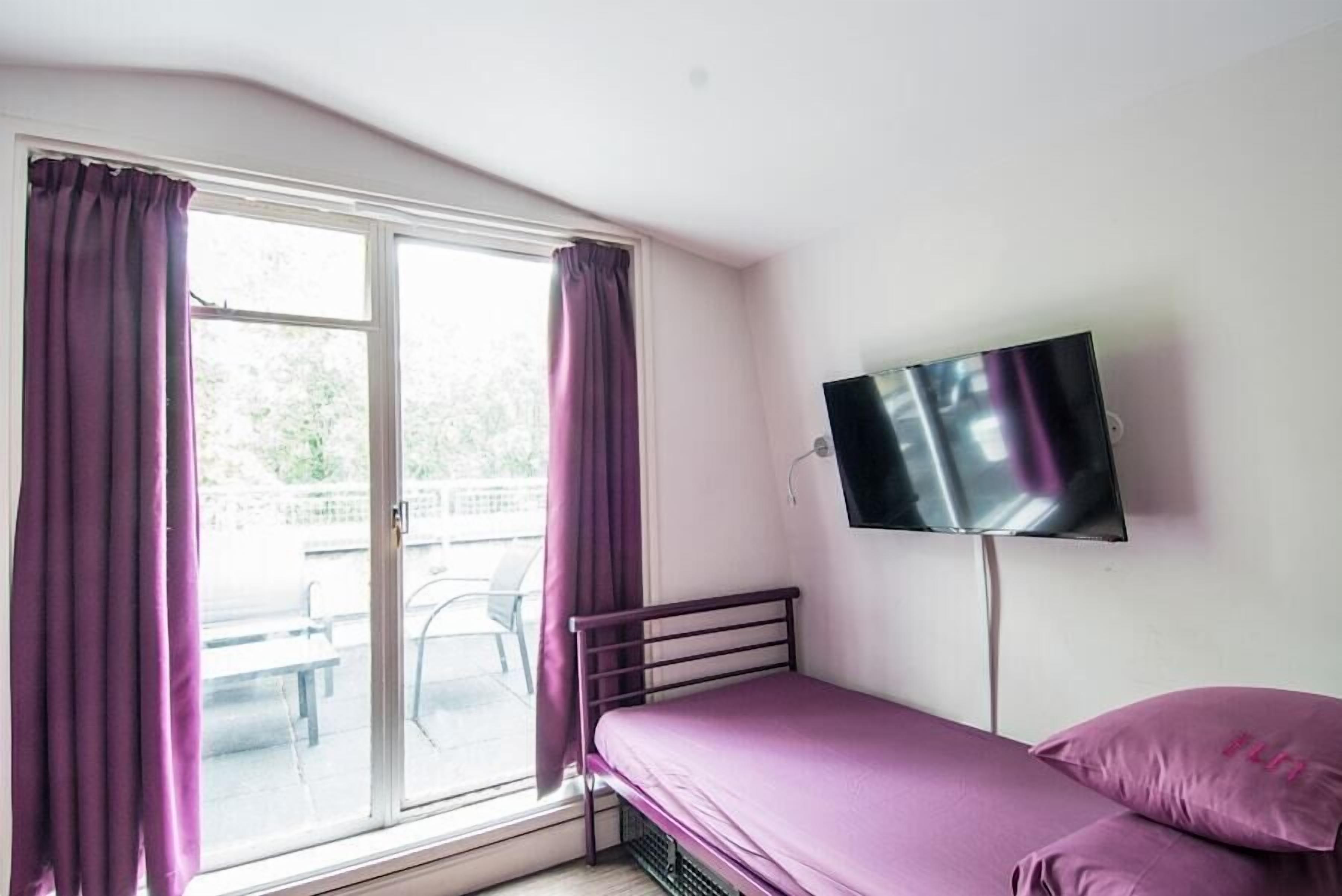 A single-bed room with a TV mounted on the wall, a window with purple curtains, and a view of an outdoor area with a table and chairs.