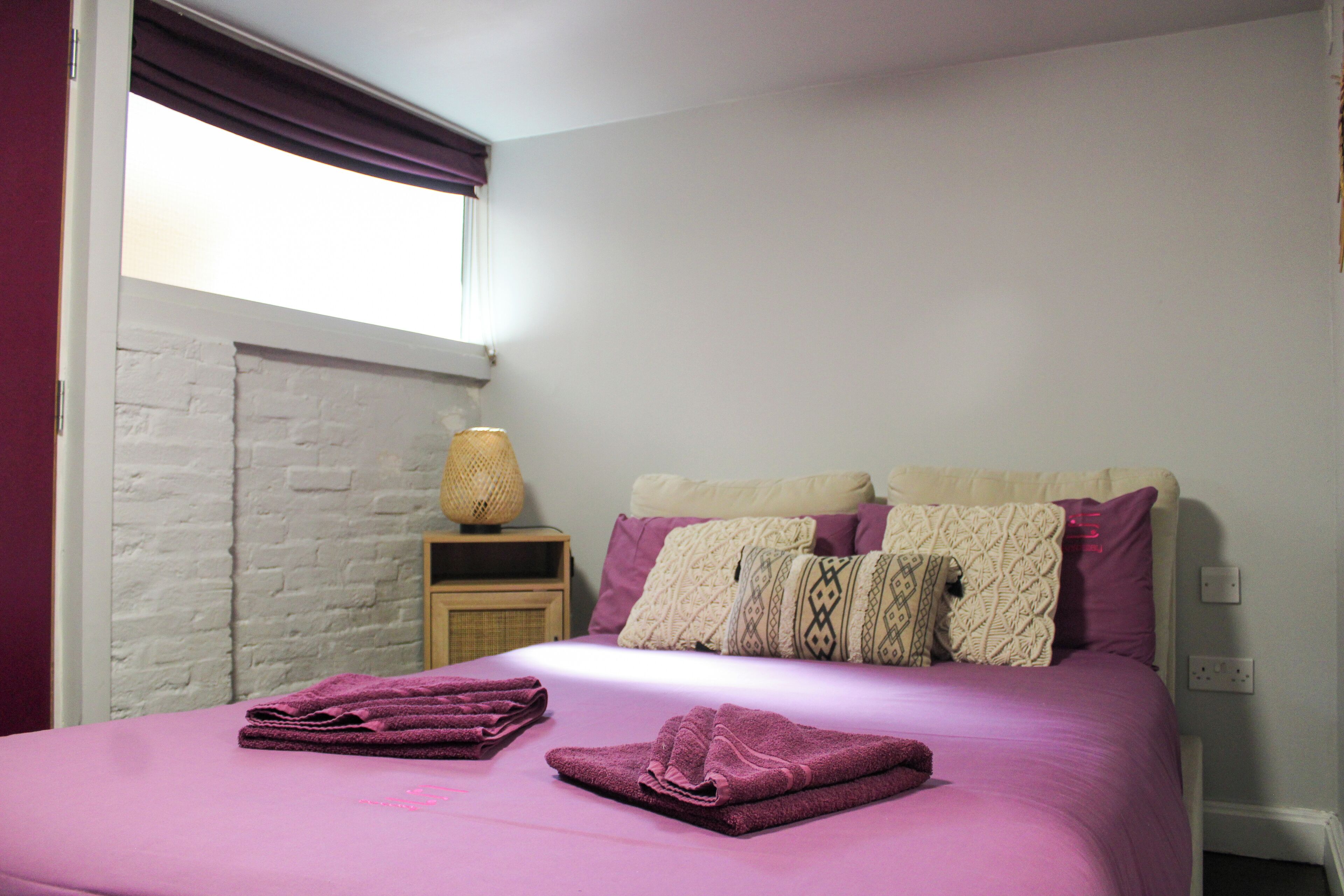A neatly made bed with purple bedding, a bedside lamp, and a white brick wall.